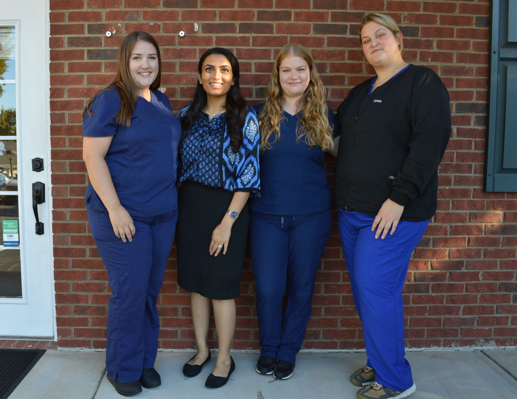Four people standing in front of a brick wall, three in blue scrubs and one in a patterned top and black skirt.