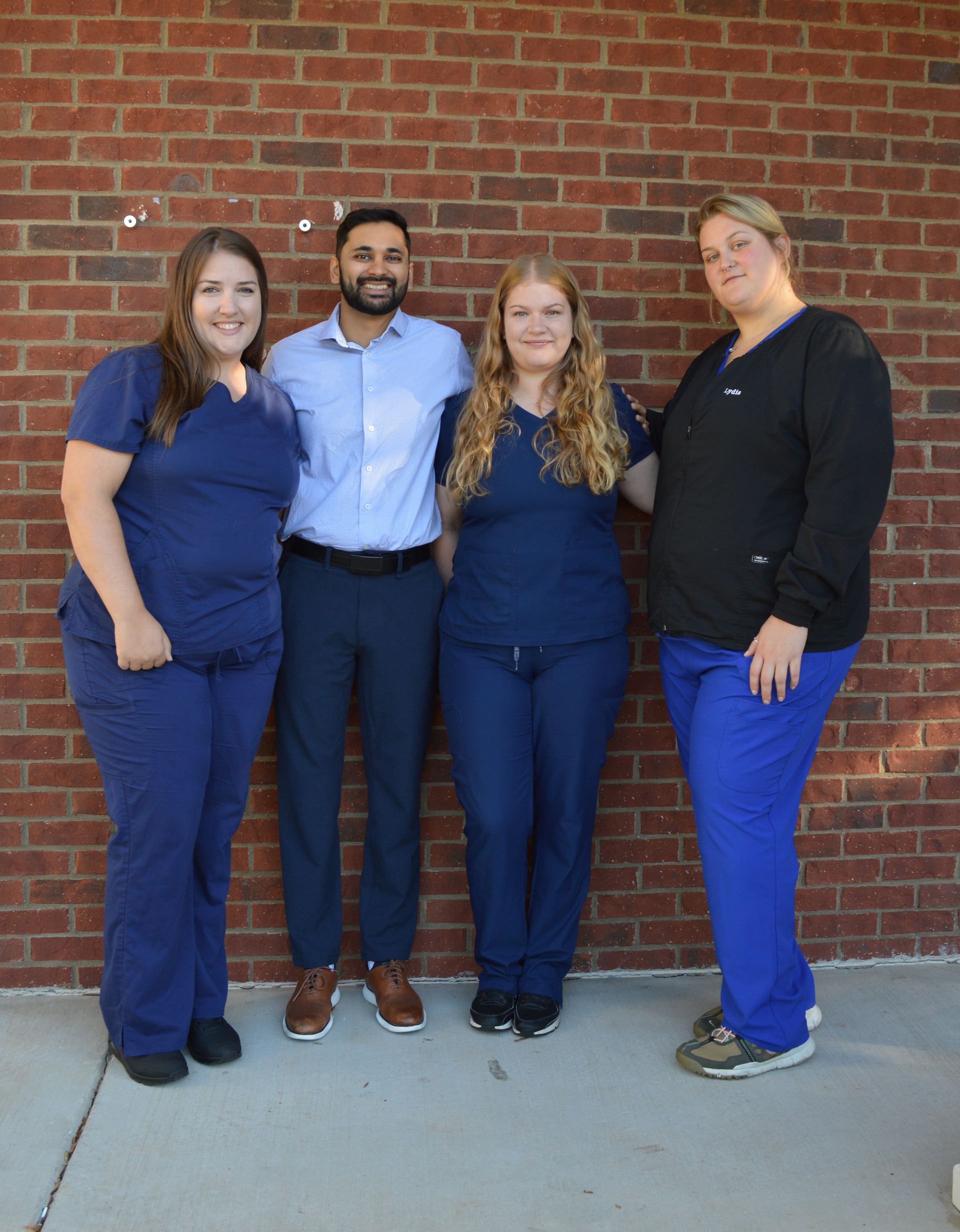 Four people stand in front of a brick wall. Three wear blue scrubs, one wears a light blue shirt and dark pants.
