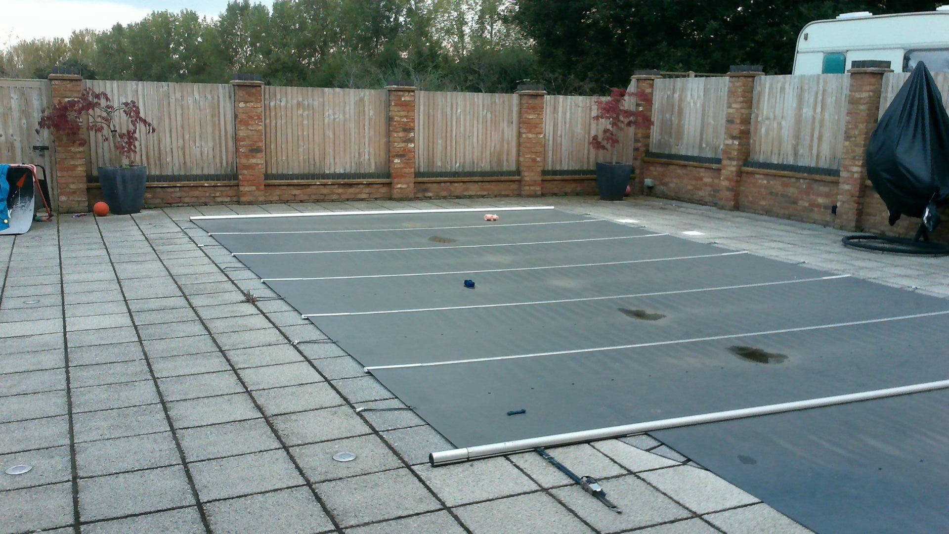Pool covered with a dark tarp on a patio, bordered by a wooden fence and brick pillars.