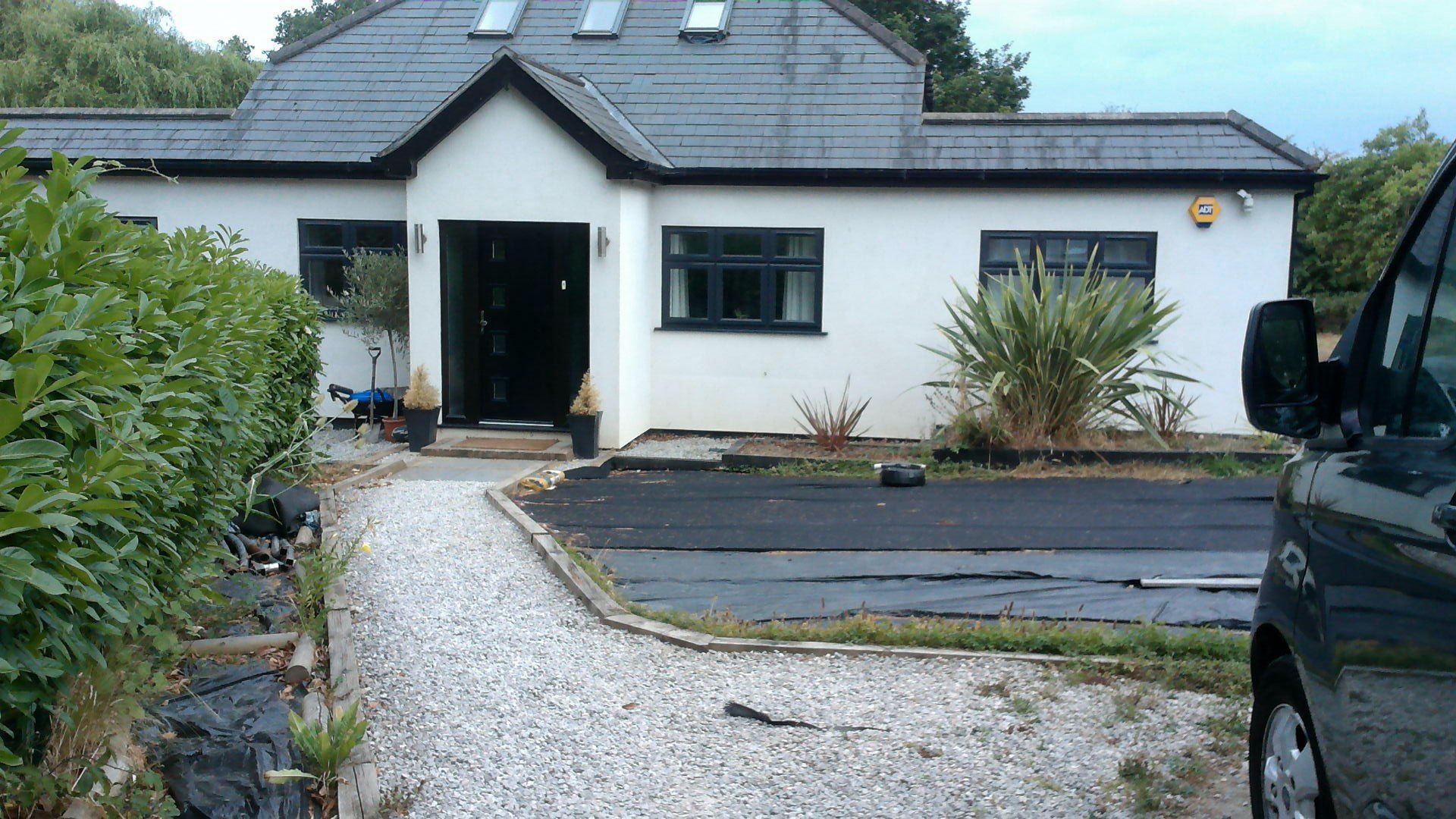 White house with black roof, windows, and door. Gray gravel path leads to the entrance. A black van is partially visible.