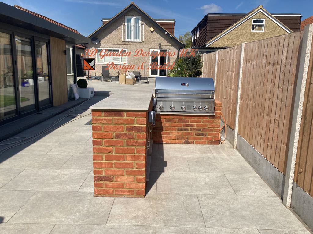 Outdoor brick grill station on paved patio, between a modern building and wooden fence.