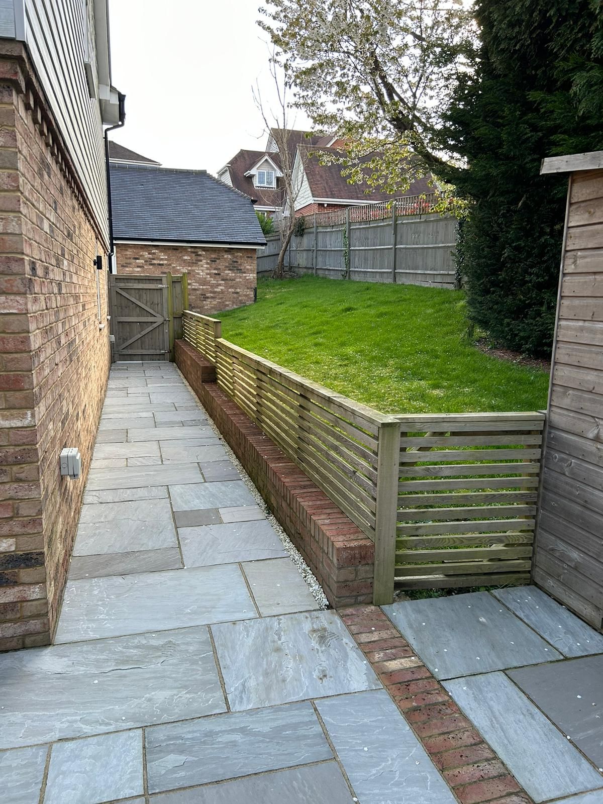 Stone walkway alongside a brick wall, leading to a grassy backyard with a wooden fence.