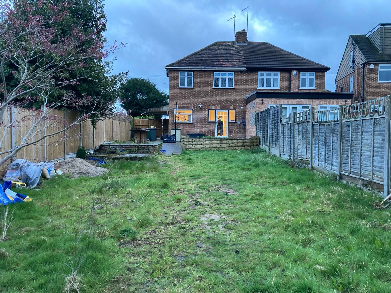 Backyard with overgrown grass, wooden fence, brick house with lit windows, and a cloudy sky.
