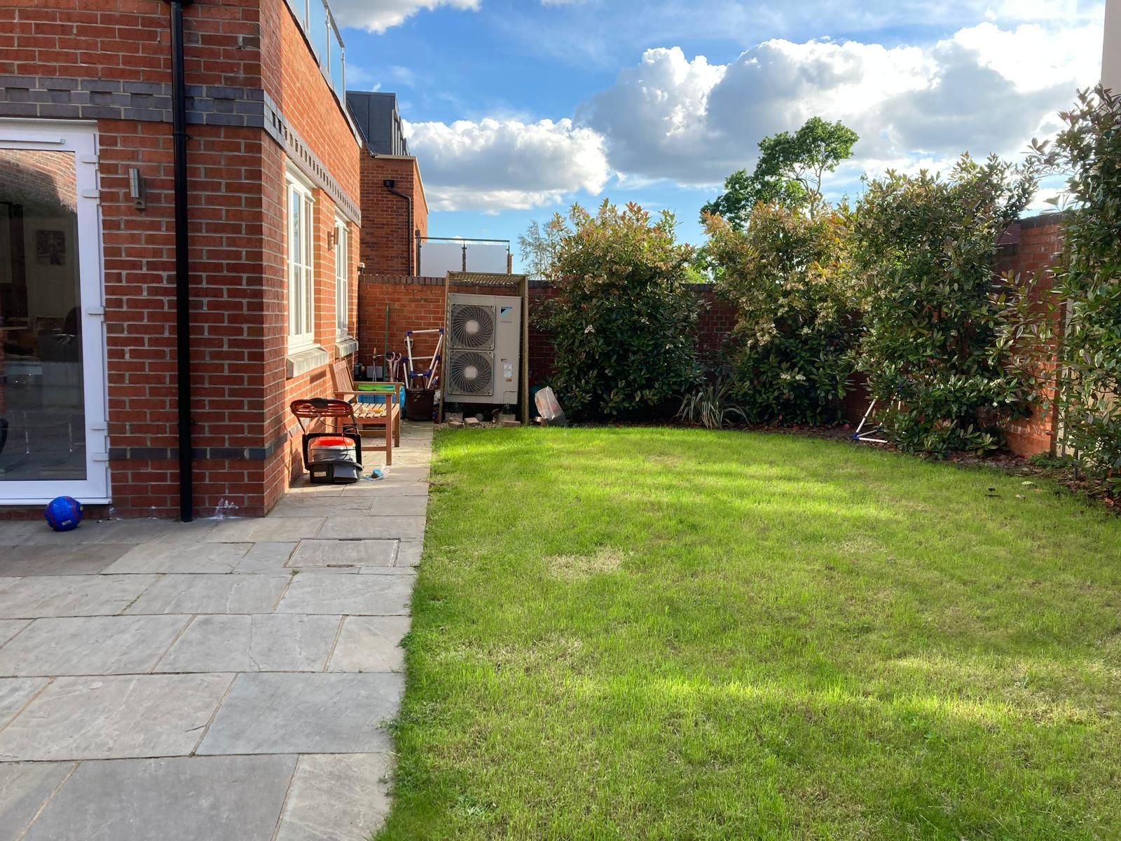 Brick house with patio and a green lawn. Cloudy sky, trees, and an air conditioning unit visible.