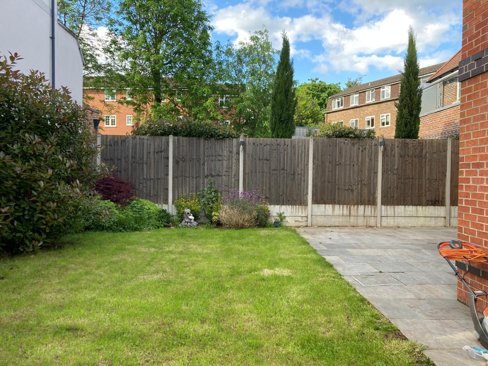 Lawn and garden with a wooden fence. Two tall, thin trees in the background. Buildings on a sunny day.