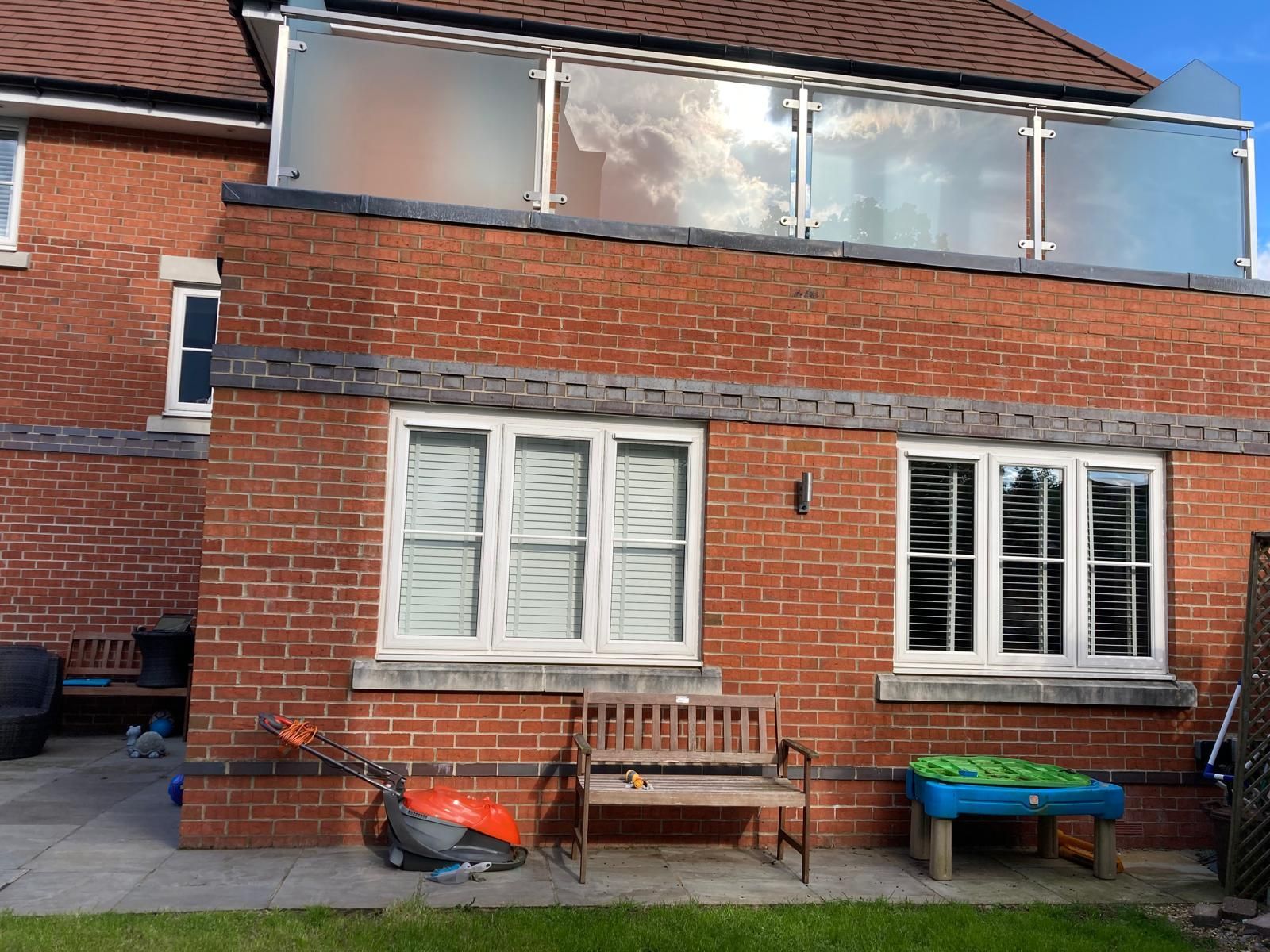 Red brick house exterior with windows, a balcony, and a small garden with a bench and tools.