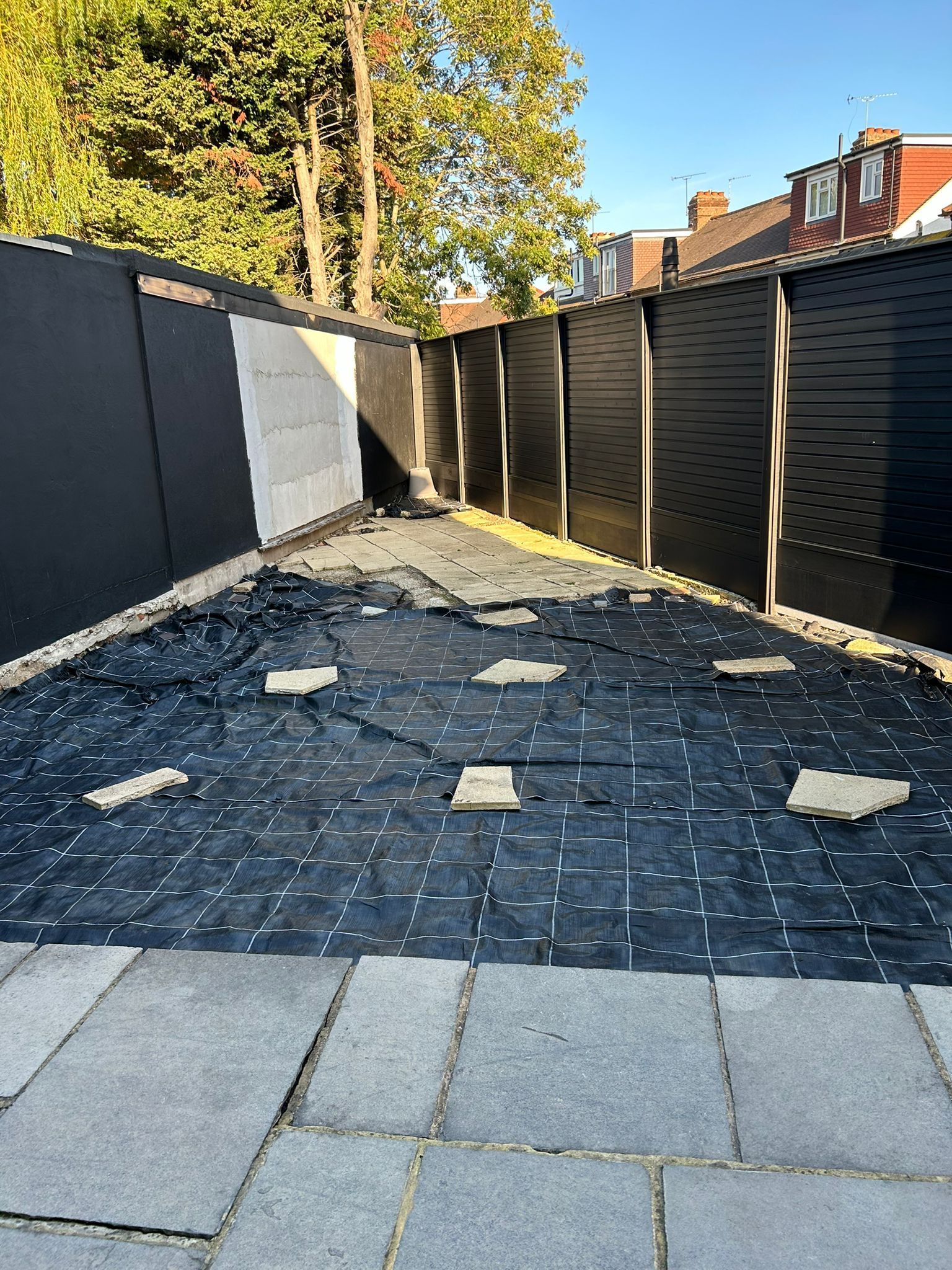Backyard with black and tan fences, torn black ground covering, and paved patio area.