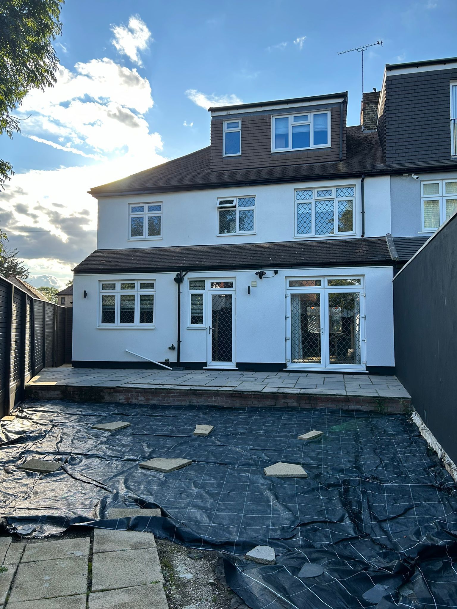 White house with black roof, multiple windows, and a paved patio in sunlight.