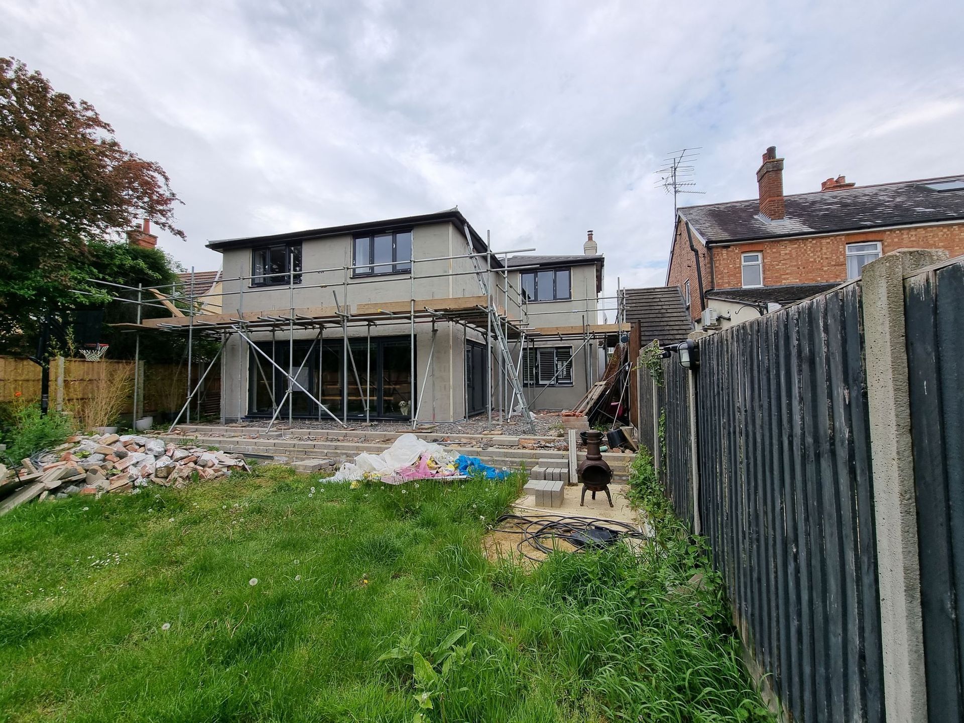 Backyard view of a house under construction; scaffolding, grey stucco, exposed beams, lawn, and a black fence.