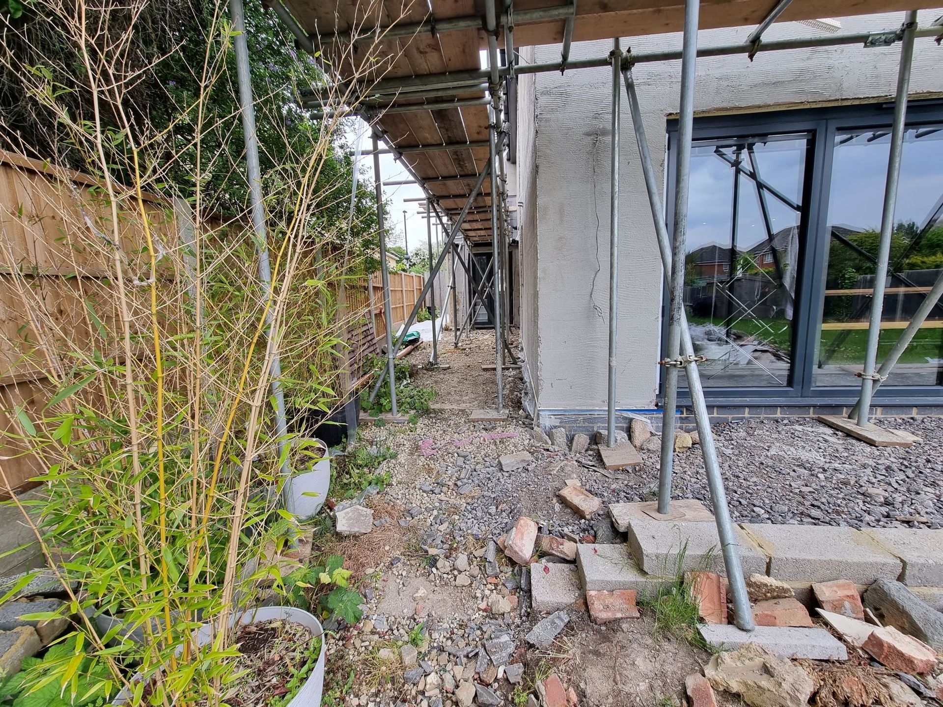 Construction site with scaffolding along the side of a building, debris on the ground, and bamboo plants in pots.