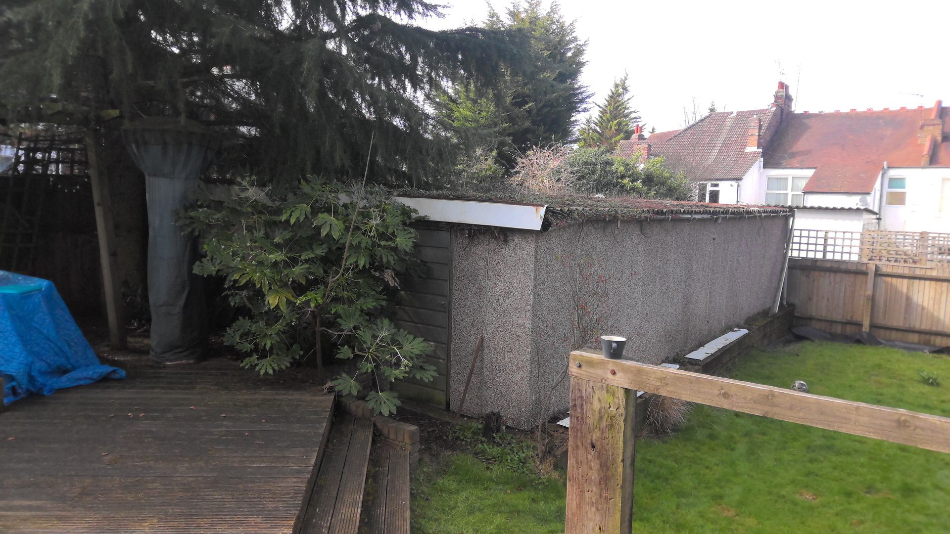 Wooden shed in a backyard, with a deck in the foreground and a fence to the right. Green lawn and trees surround it.