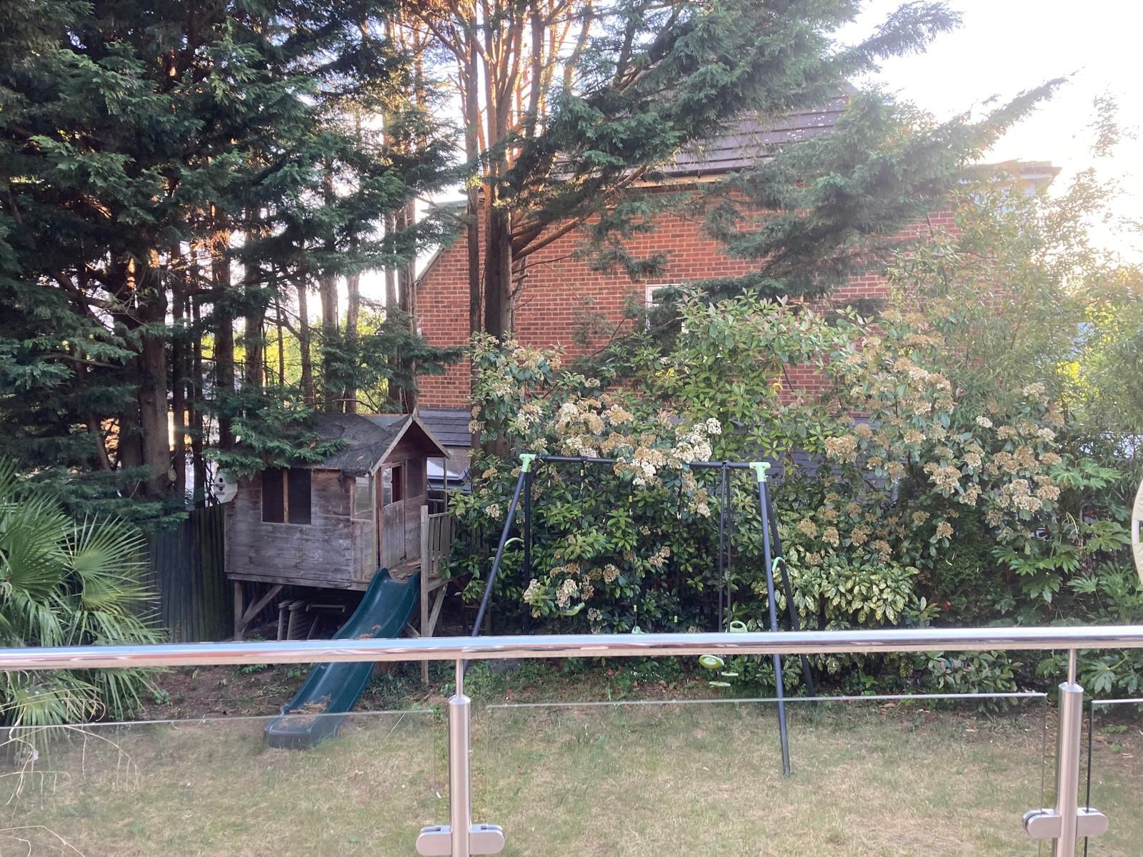 Backyard scene with a two-story brick house, a wooden playhouse, swings, and lush trees.
