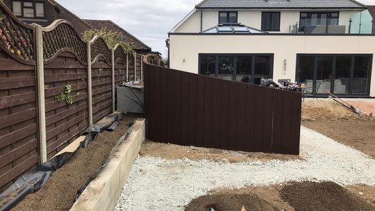 Brown fence with gravel path and backyard landscaping. Modern house in background.