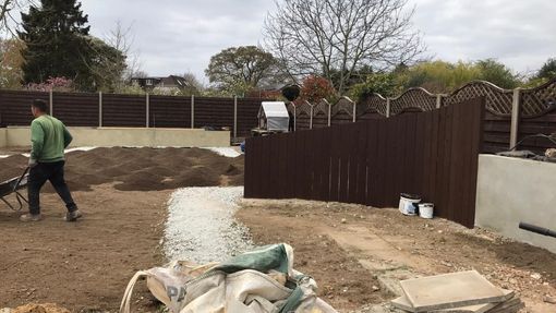 Man in a green hoodie pushing a wheelbarrow in a yard under construction with brown fence and gravel pathway.