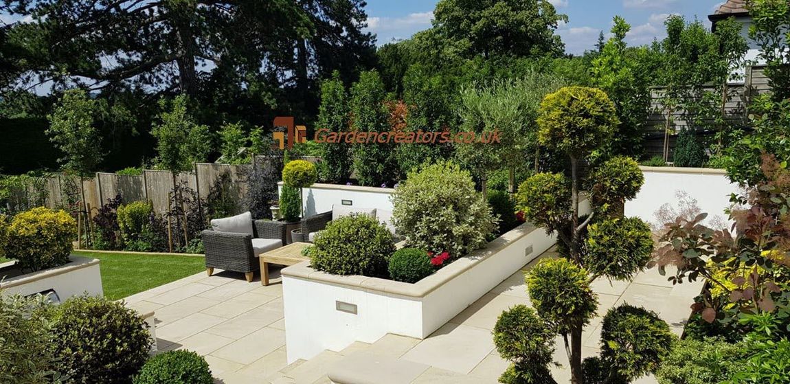 Sunny garden with raised white planters, colorful flowers, shrubs, and a stone patio under trees