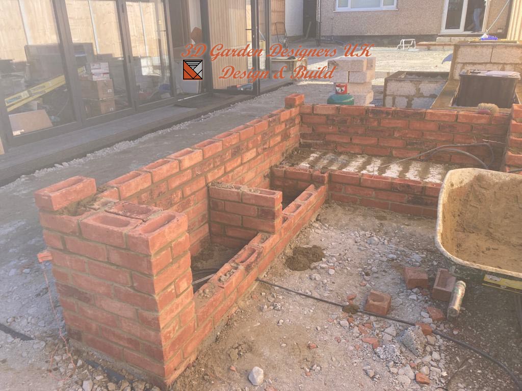 Brickwork construction in progress outdoors, forming a low wall structure. Red bricks are being laid.
