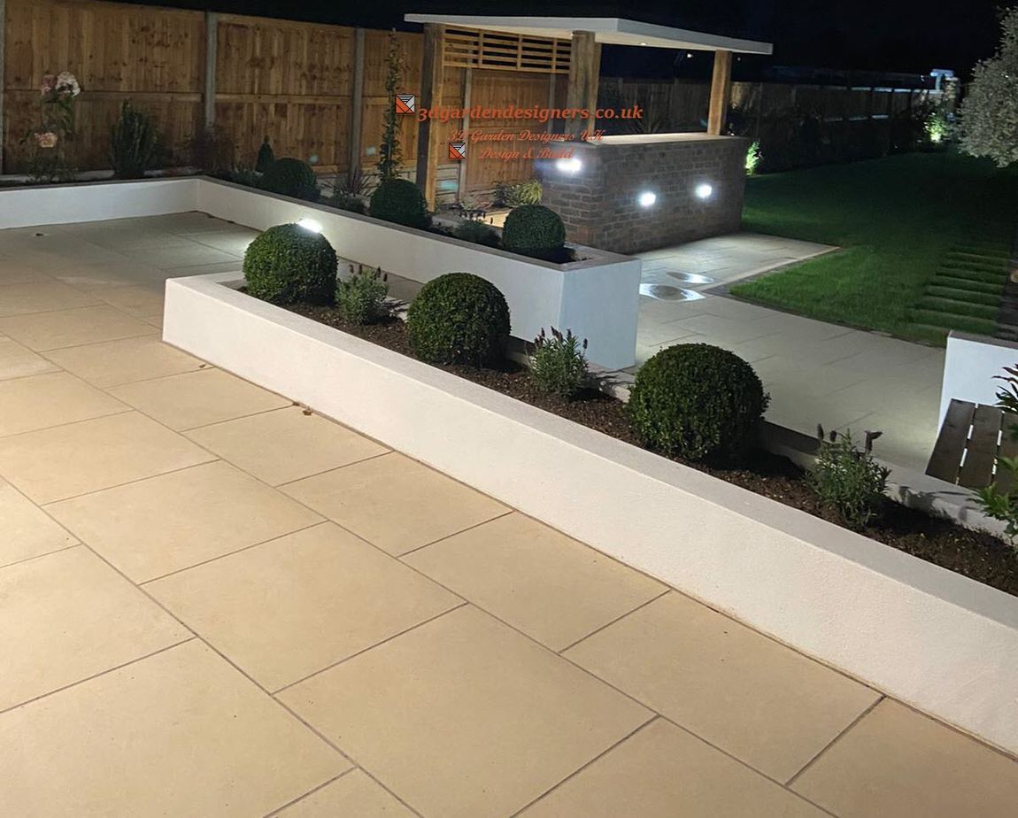 Nighttime view of a modern backyard patio with illuminated stone walls, plants, and a bar area with lights.