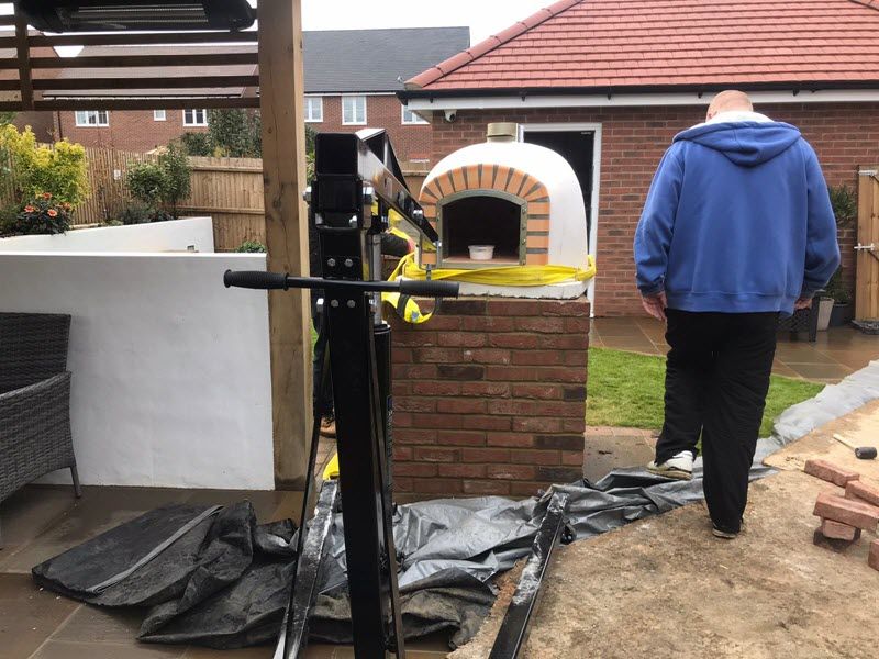 Man walks toward a brick pizza oven under construction, outdoors. Yellow tape surrounds the oven, red brick base.