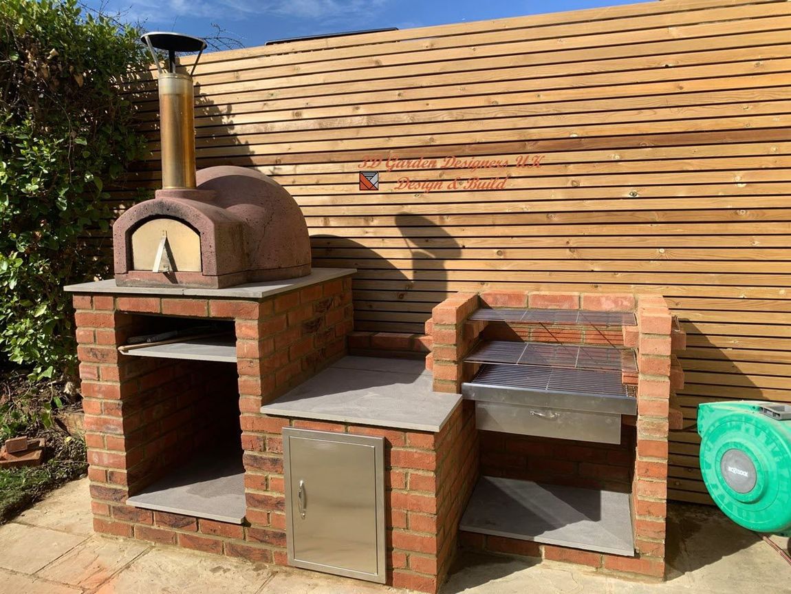 A brick outdoor kitchen featuring a pizza oven with a metal chimney and a charcoal grill, set against a wooden fence.