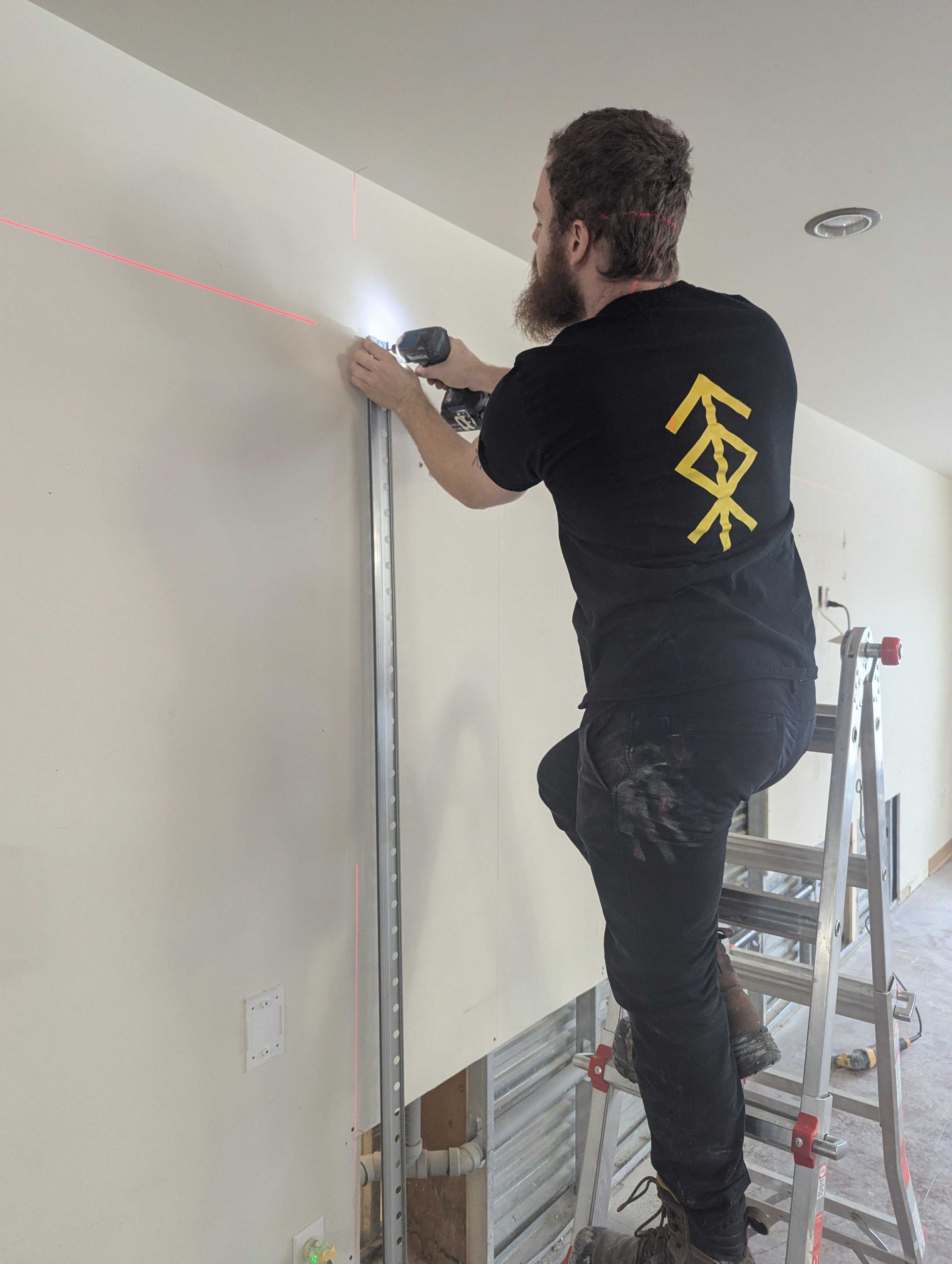 Un homme, juché sur une échelle, installe un montant métallique à l'aide d'un niveau laser. Travail en intérieur, sur un mur.