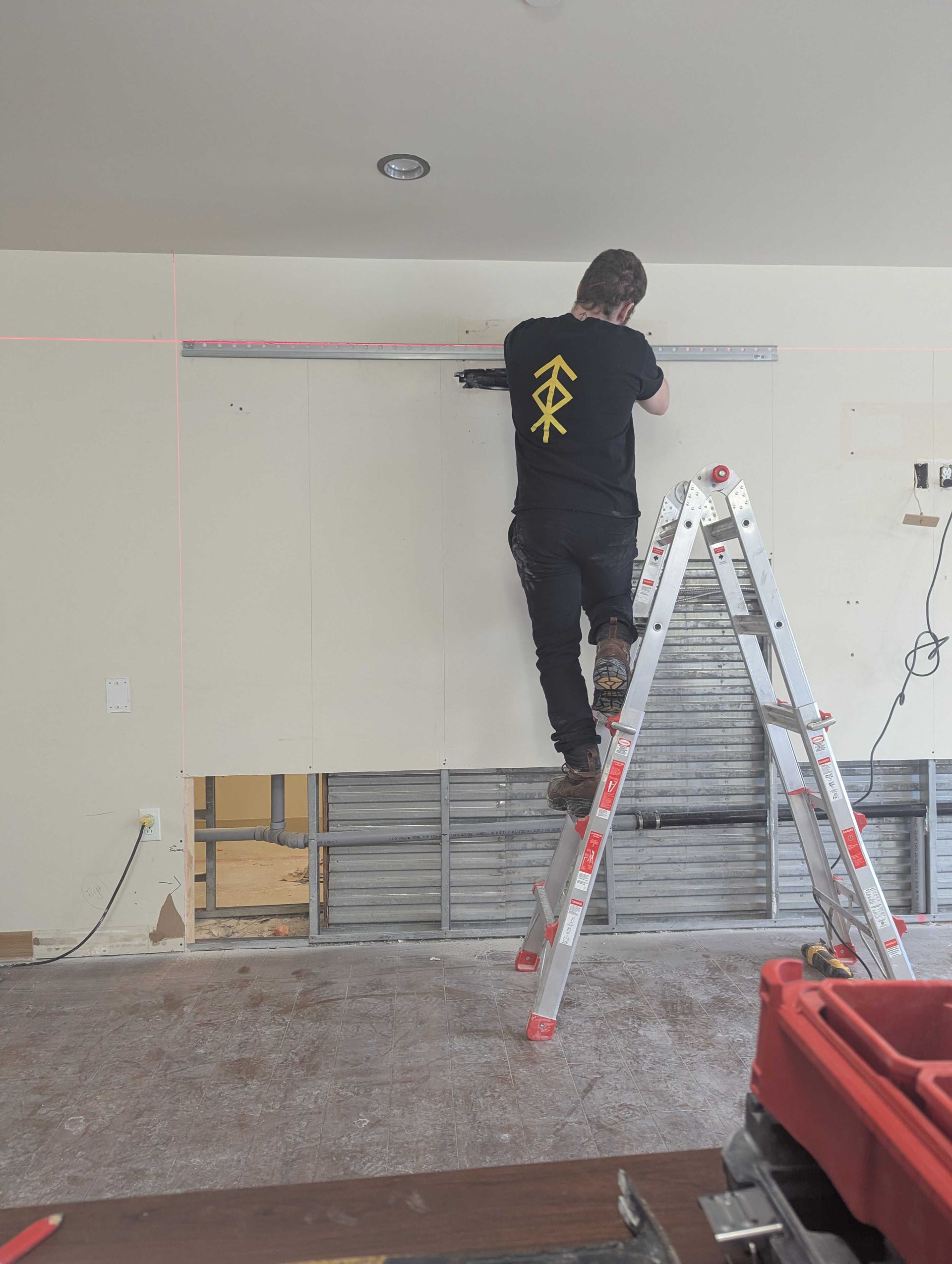 Une personne sur un escabeau mesure un mur avec un niveau laser dans une pièce en construction.