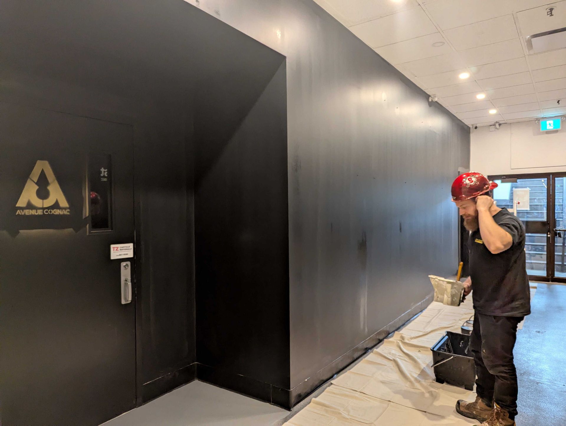 Une personne portant un casque de chantier peint un mur sombre dans un couloir où se trouve une porte noire à gauche.