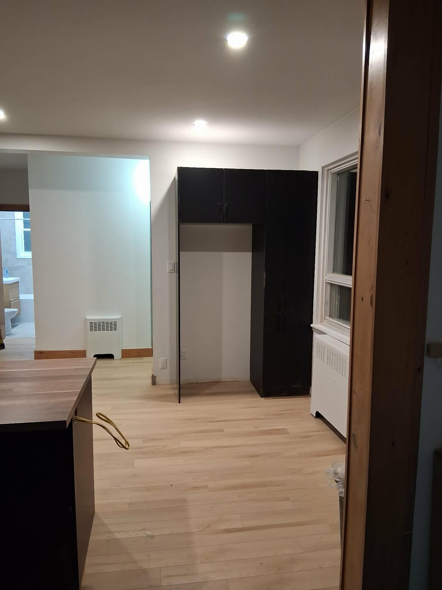 Vue intérieure d'une cuisine avec parquet clair, placards noirs, murs blancs et une fenêtre.
