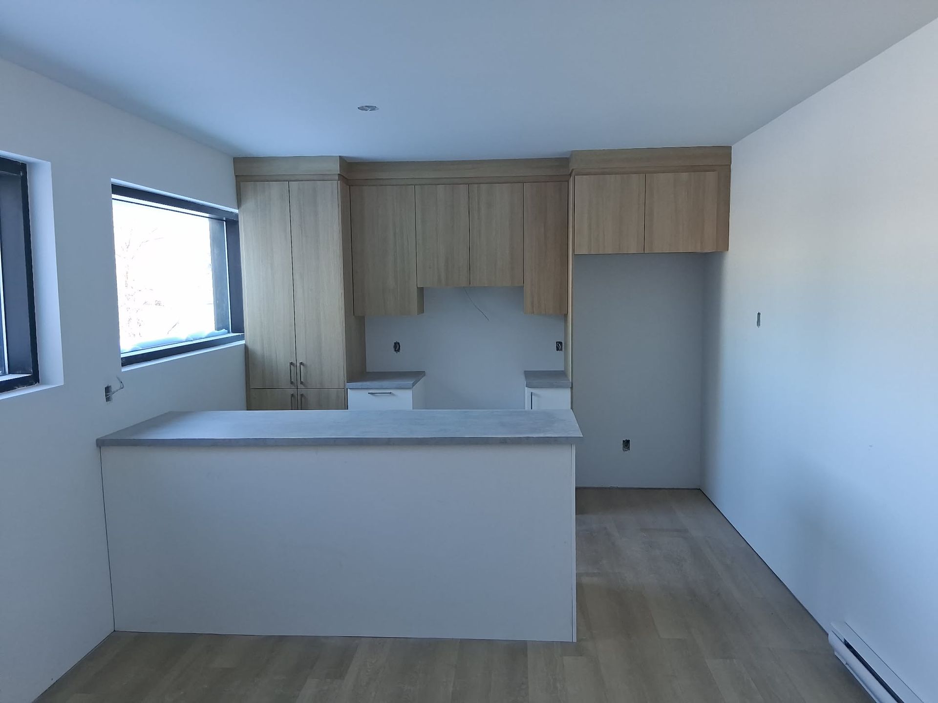 Cuisine vide avec placards en bois clair, plans de travail gris et une fenêtre.