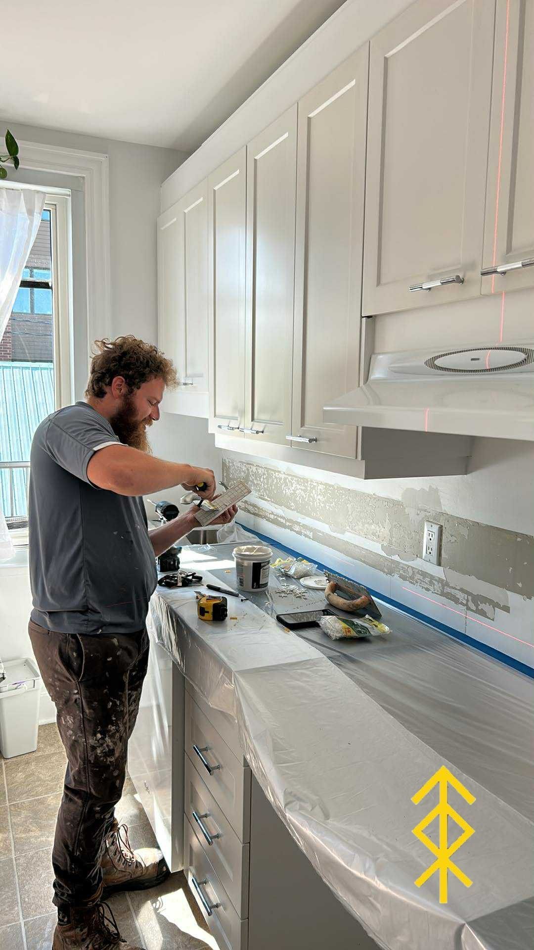 Personne posant du carrelage pour une crédence de cuisine. Placards blancs, plan de travail recouvert de plastique, lumière naturelle.