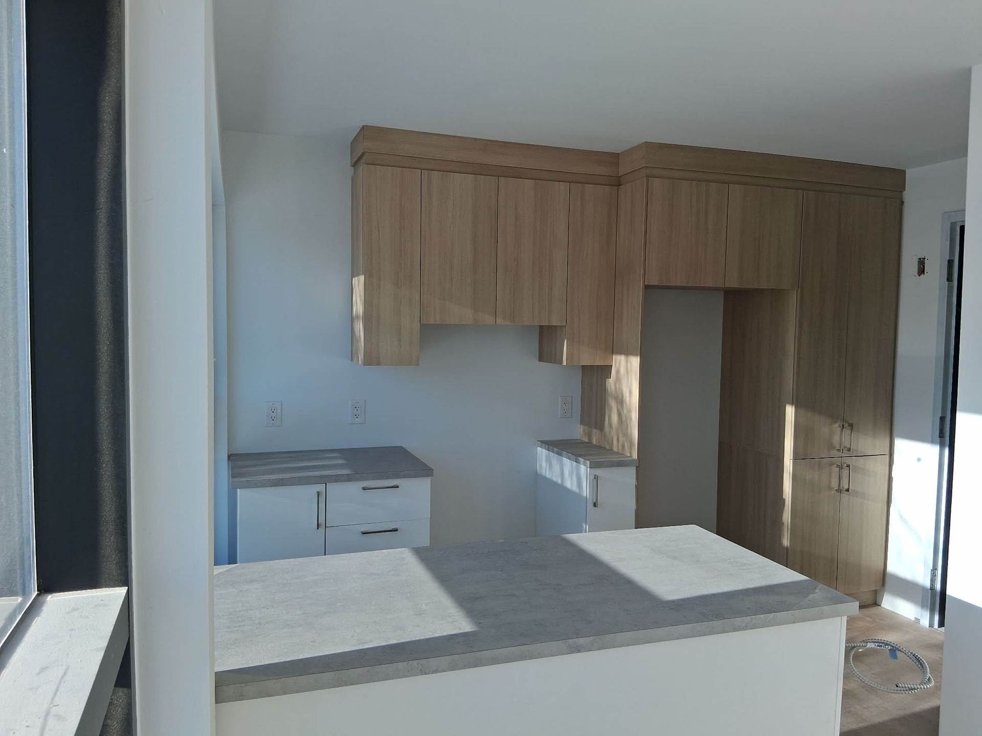 Cuisine en construction avec armoires en bois clair, comptoirs blancs et îlot central blanc.