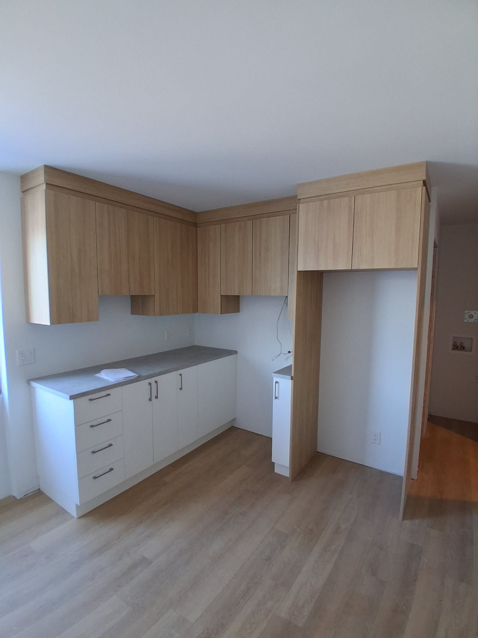Cuisine avec placards en bois clair et meubles bas blancs, comptoir et niche pour réfrigérateur vide.