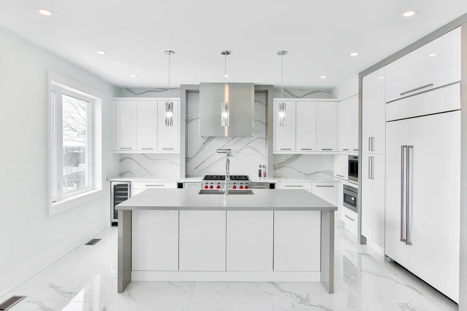 Cuisine blanche moderne avec îlot central, éléments en marbre, appareils électroménagers en acier inoxydable et luminaires suspendus.