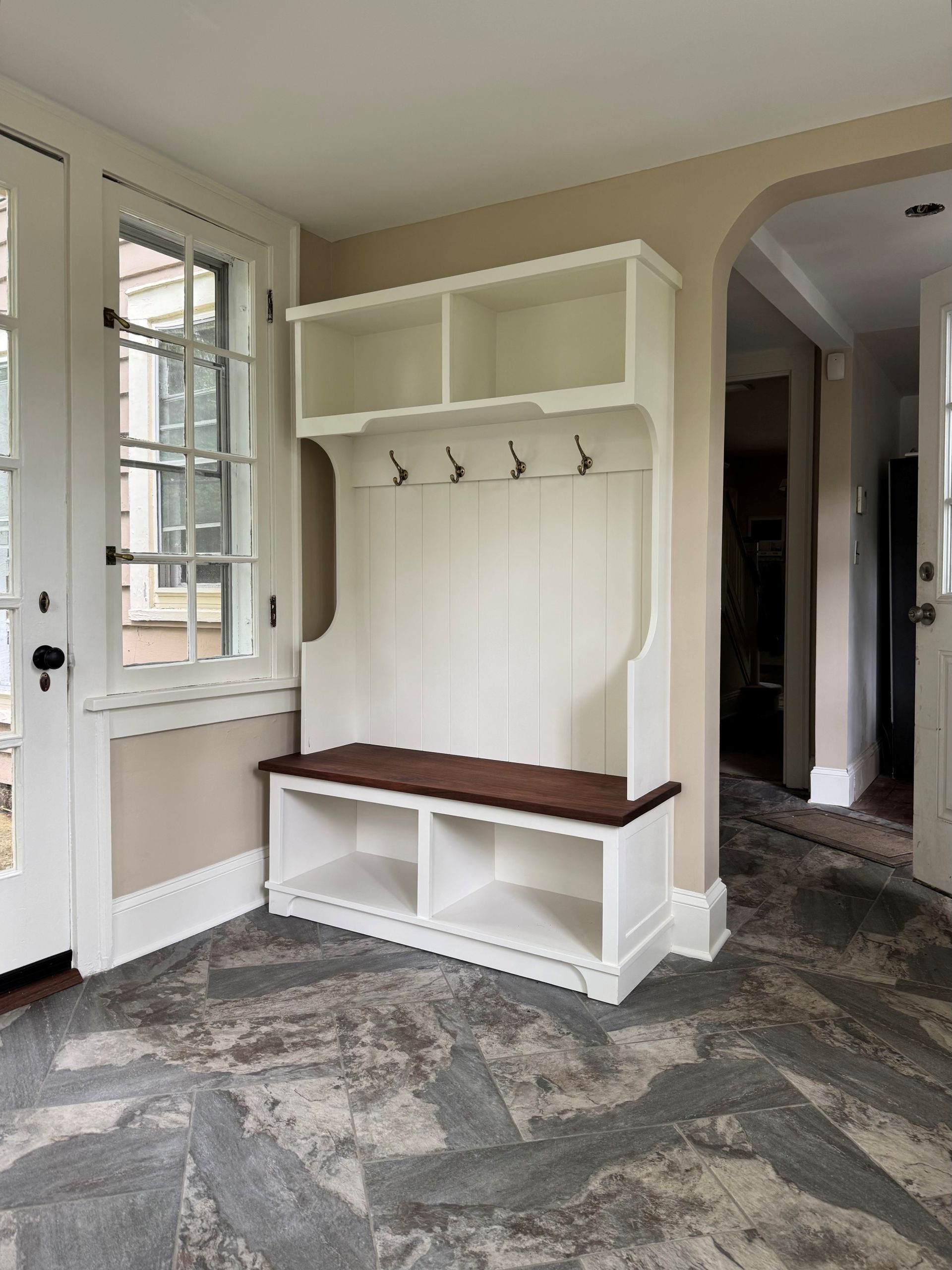 A white mudroom hall tree with a dark wood bench sits in a room with a window and gray patterned flooring.
