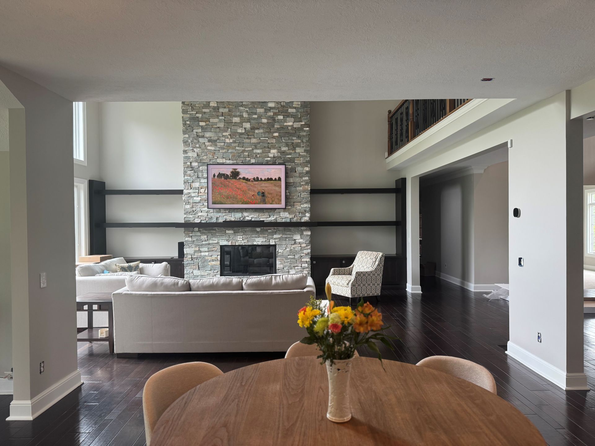A dining table with flowers in the foreground, facing a living room with a stone fireplace, floating shelves, and sofa.