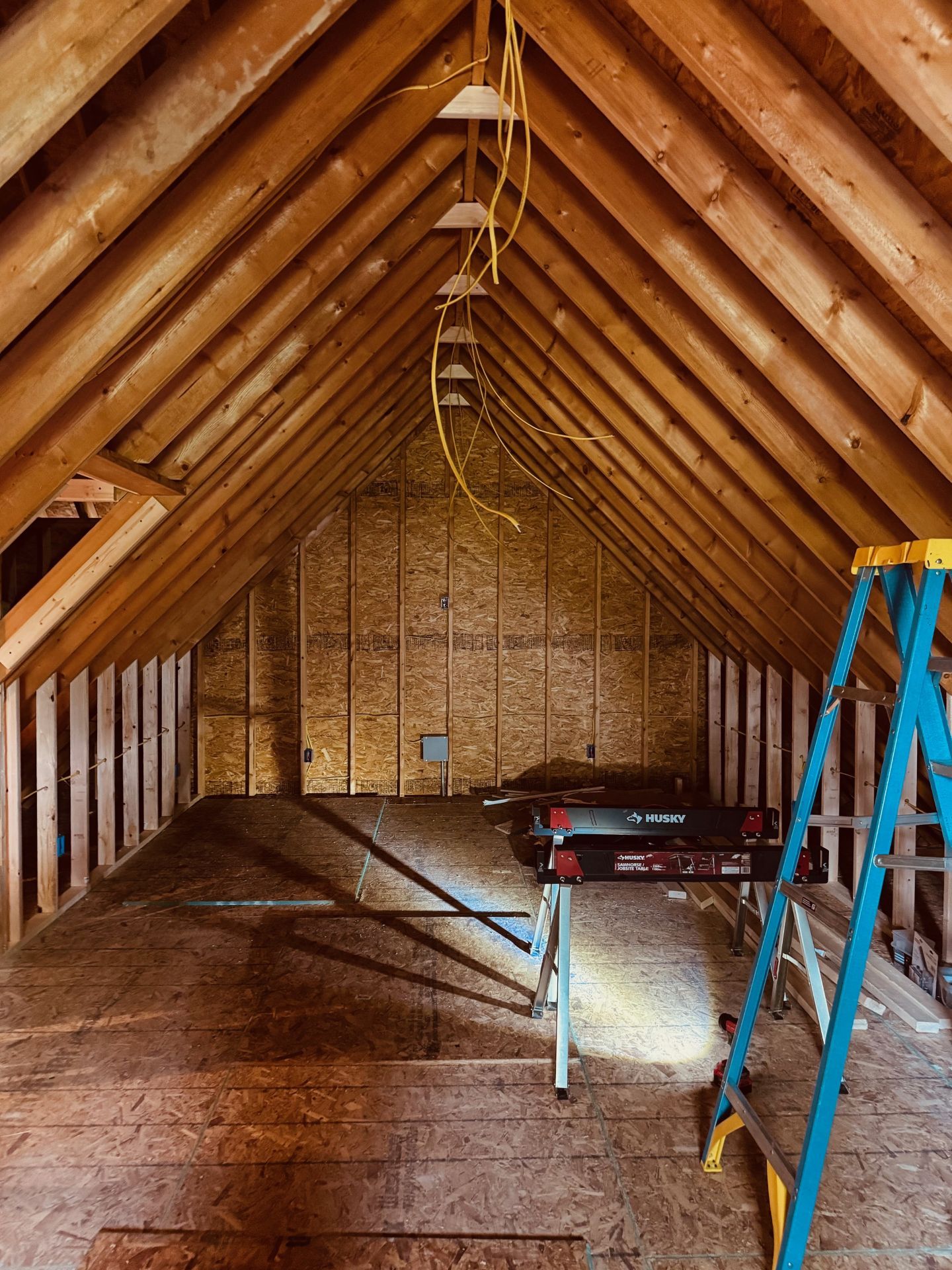 An unfinished attic with exposed wooden rafters, plywood floors, and walls, featuring a blue stepladder and electrical wire.