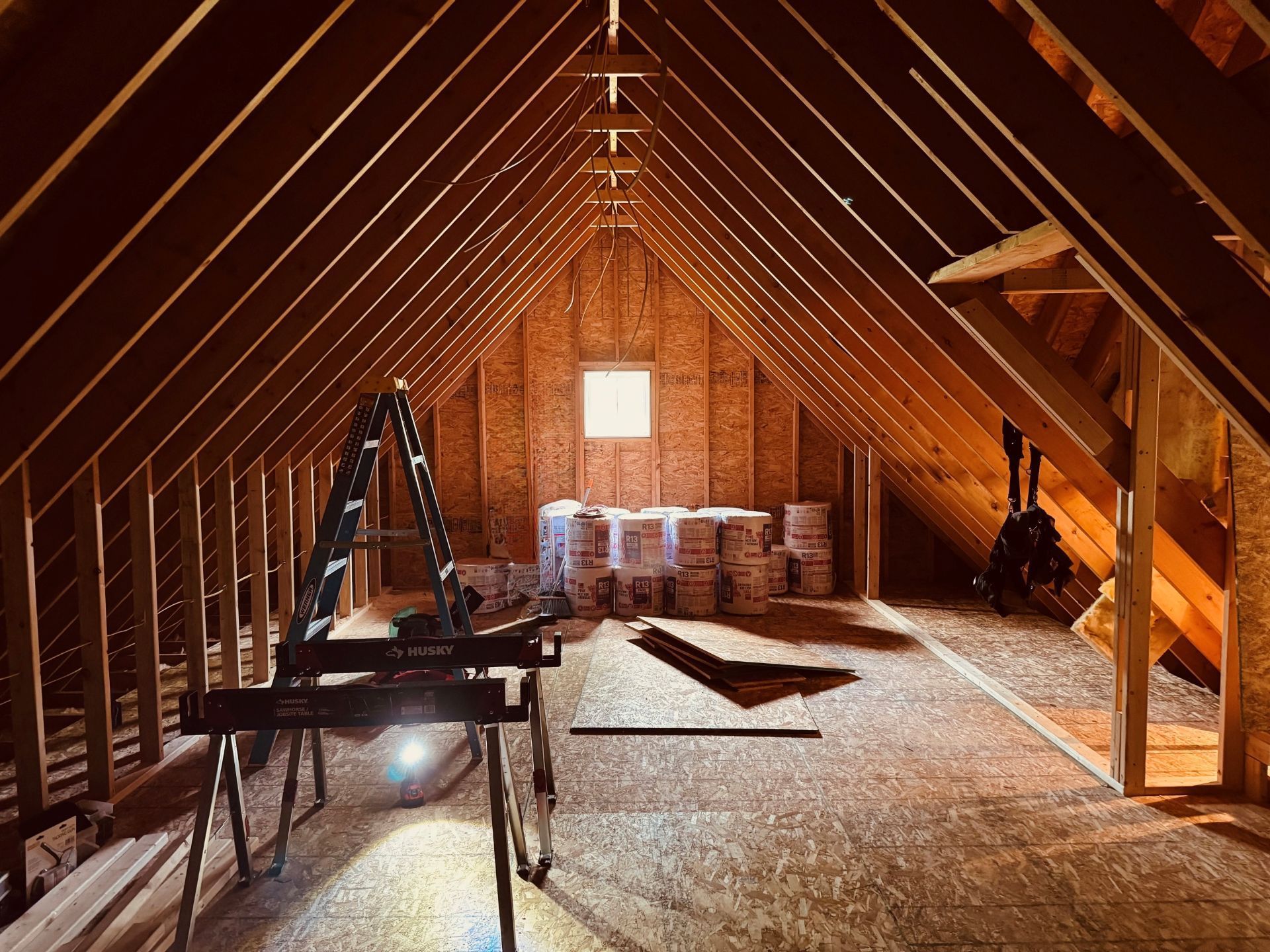 Unfinished attic space with wooden rafters, a central window, and stacks of insulation materials on a plywood floor.