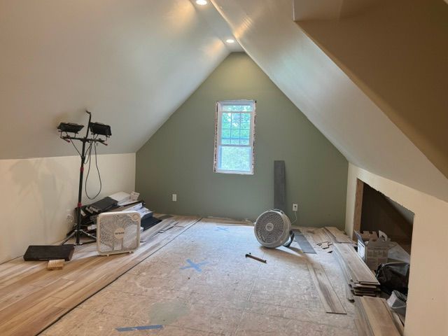 Attic room under construction with sloped walls, a window, wood flooring planks, a work light, and two fans.