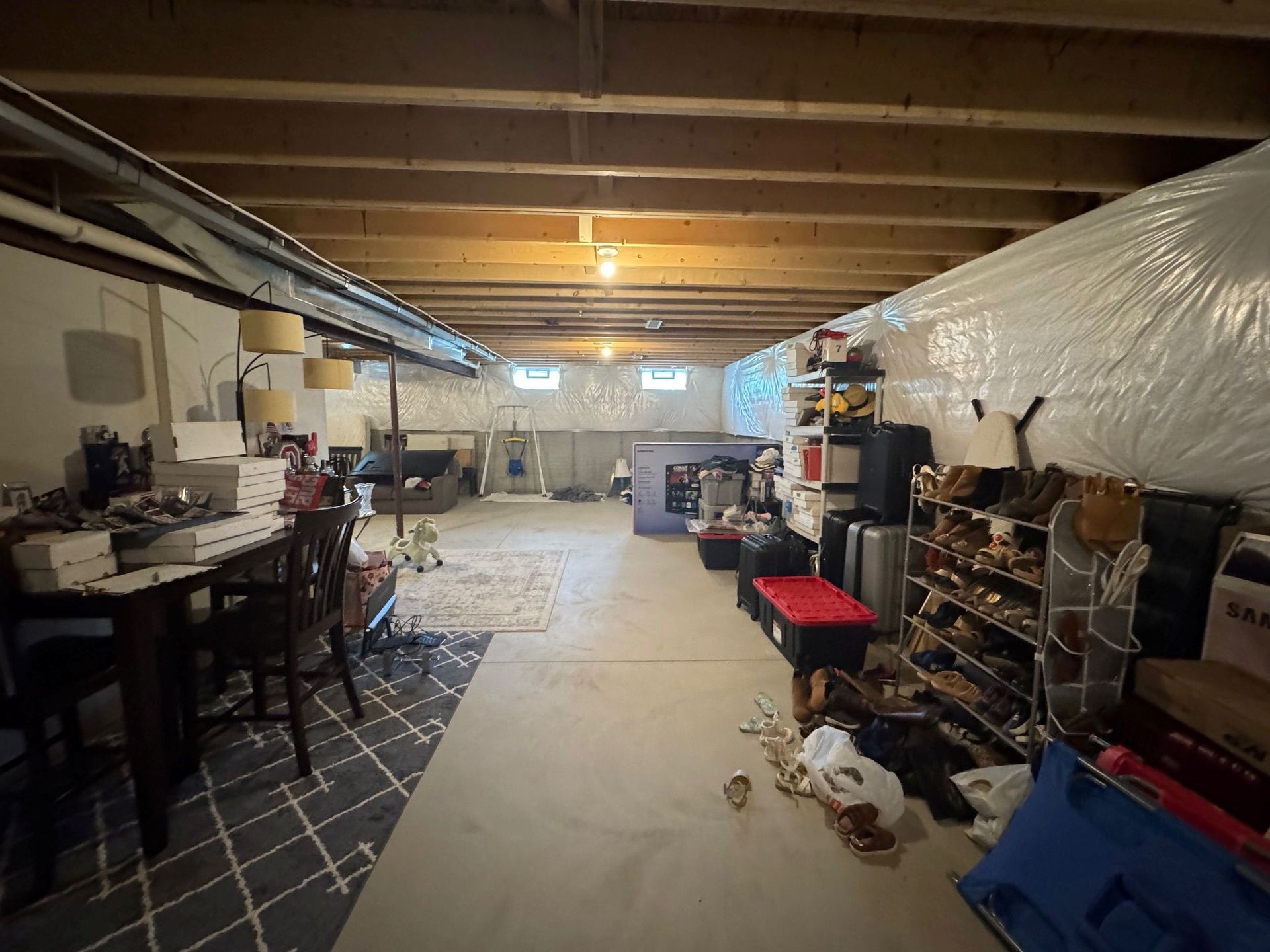 A cluttered unfinished basement with exposed wooden joists, concrete flooring, a desk area, and a shoe storage rack.