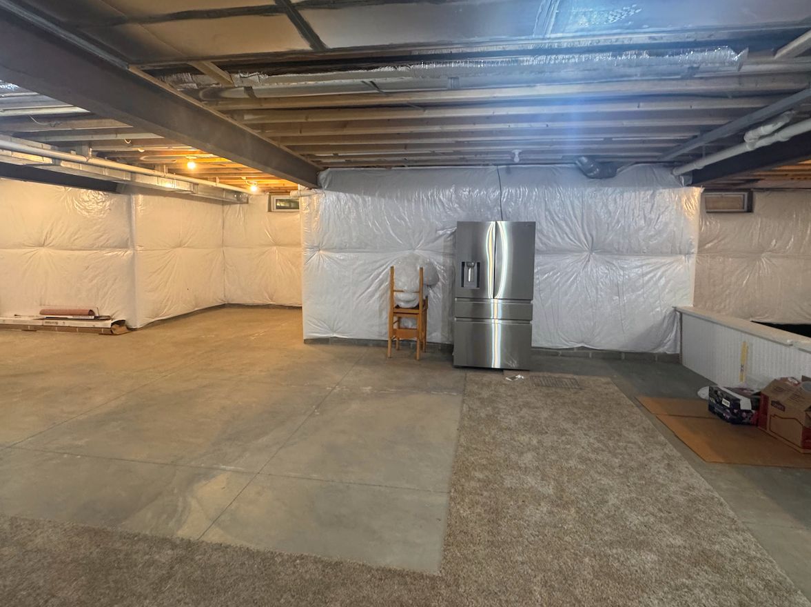 A basement with unfinished wooden ceiling beams, white insulated walls, concrete floors, and a stainless steel fridge.
