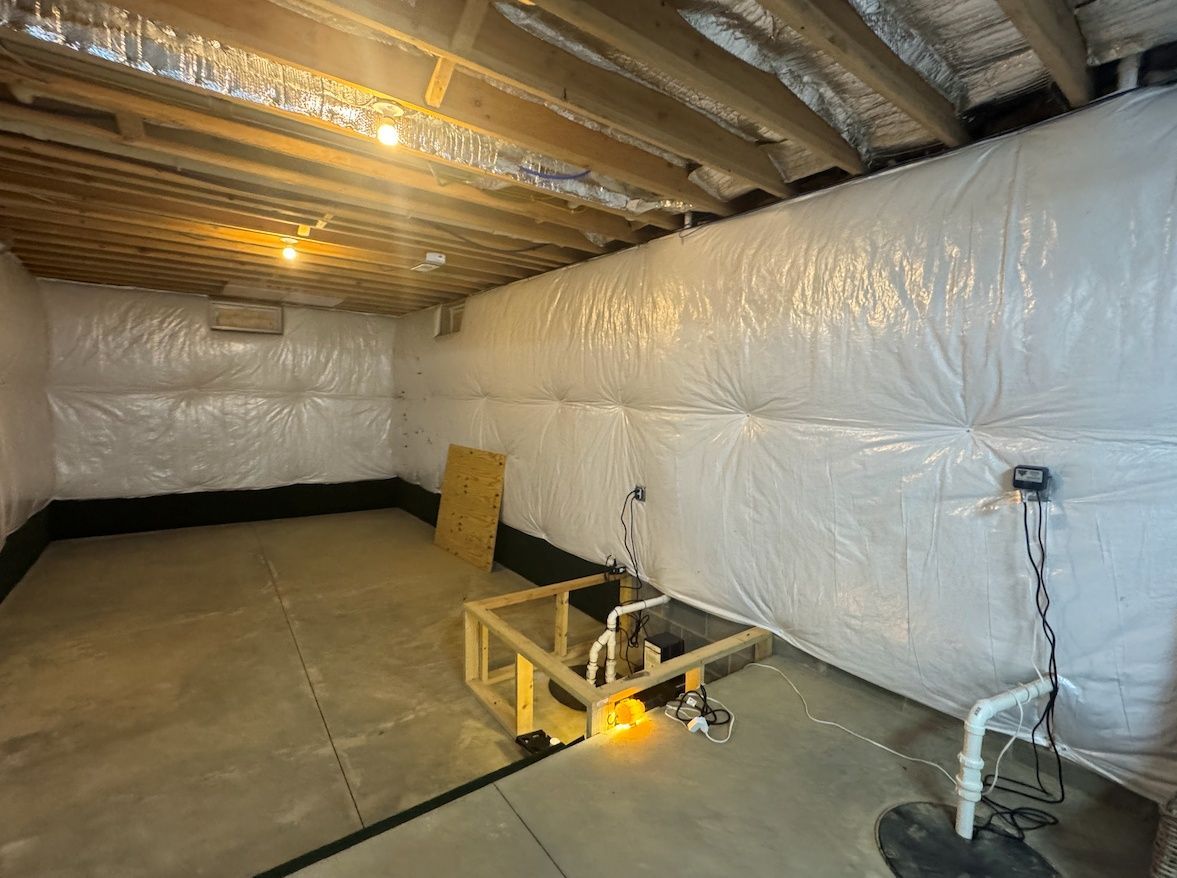 Unfinished basement with white vapor barrier walls, exposed wooden ceiling joists, a wooden frame, and a sump pump.