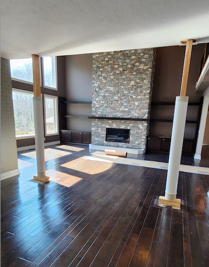 A living room with dark hardwood floors, two support columns, and a large stone fireplace with built-in shelves.