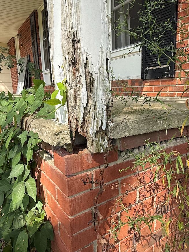 A rotted white wooden porch support column rests on a red brick foundation, with overgrown greenery nearby.