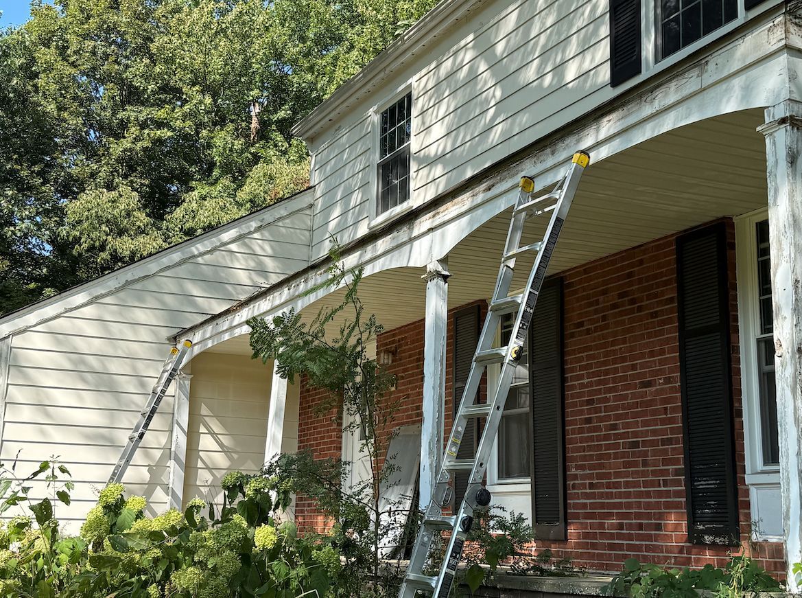 An extension ladder leans against the white siding and porch roof of a two-story brick and siding house.