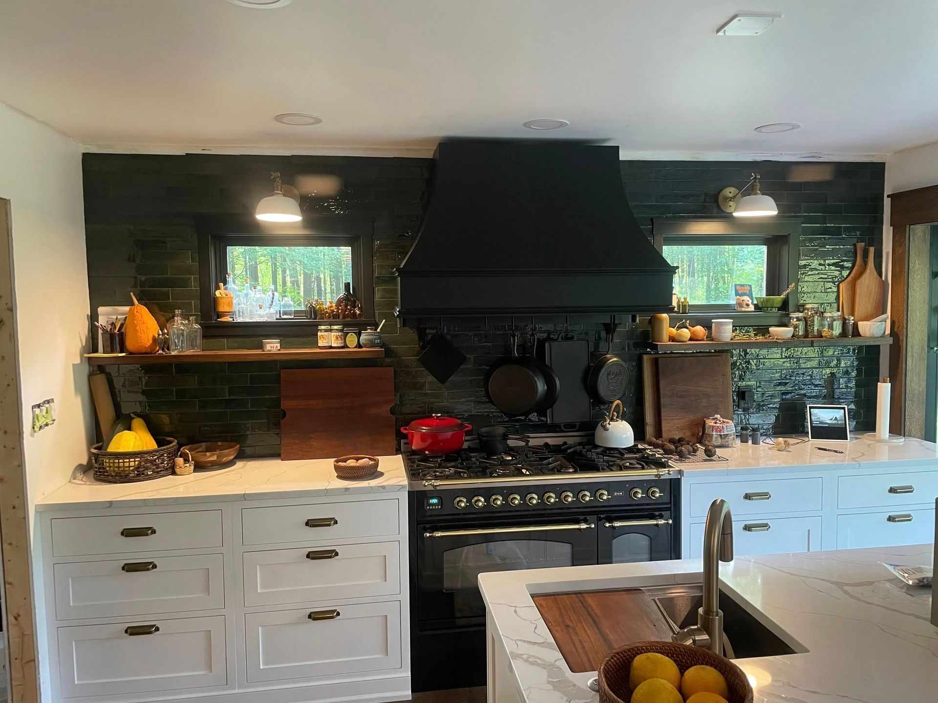 A kitchen featuring white cabinets, a black stove and hood, and dark tiled walls with two windows and wooden shelves.