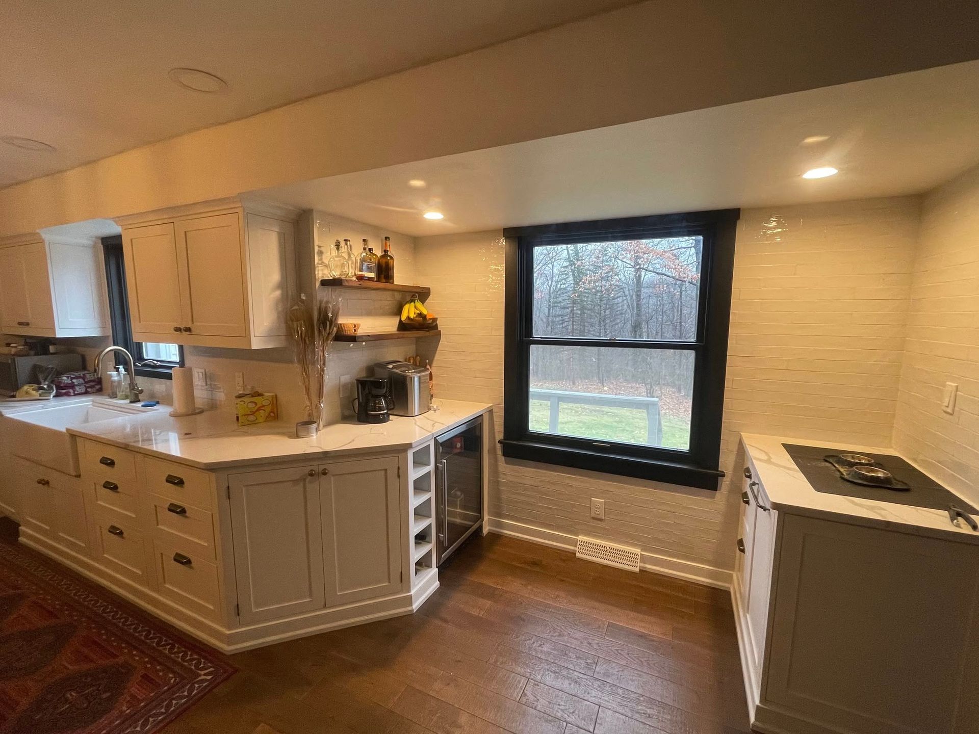 A modern kitchen with off-white cabinets, dark countertops, a farmhouse sink, and a black-framed window overlooking trees.