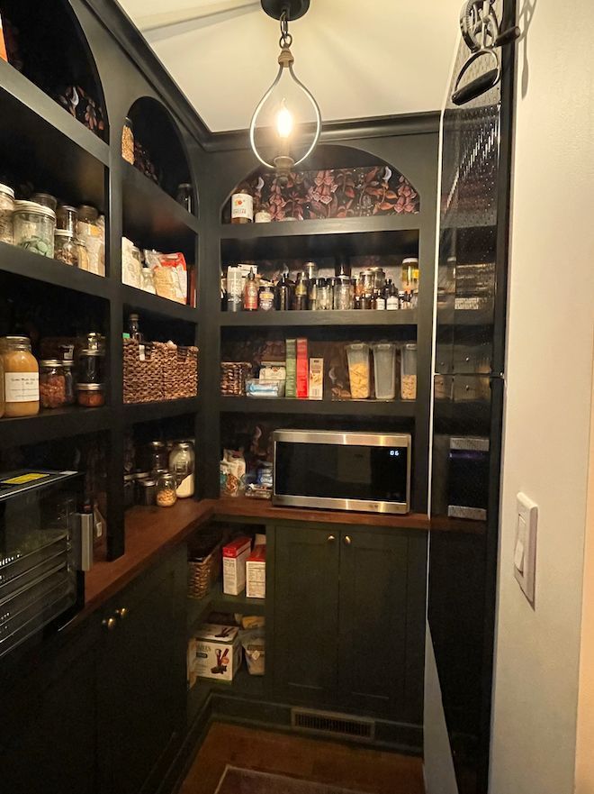 A walk-in pantry with black shelving, a microwave on the counter, patterned wallpaper, and a pendant light.