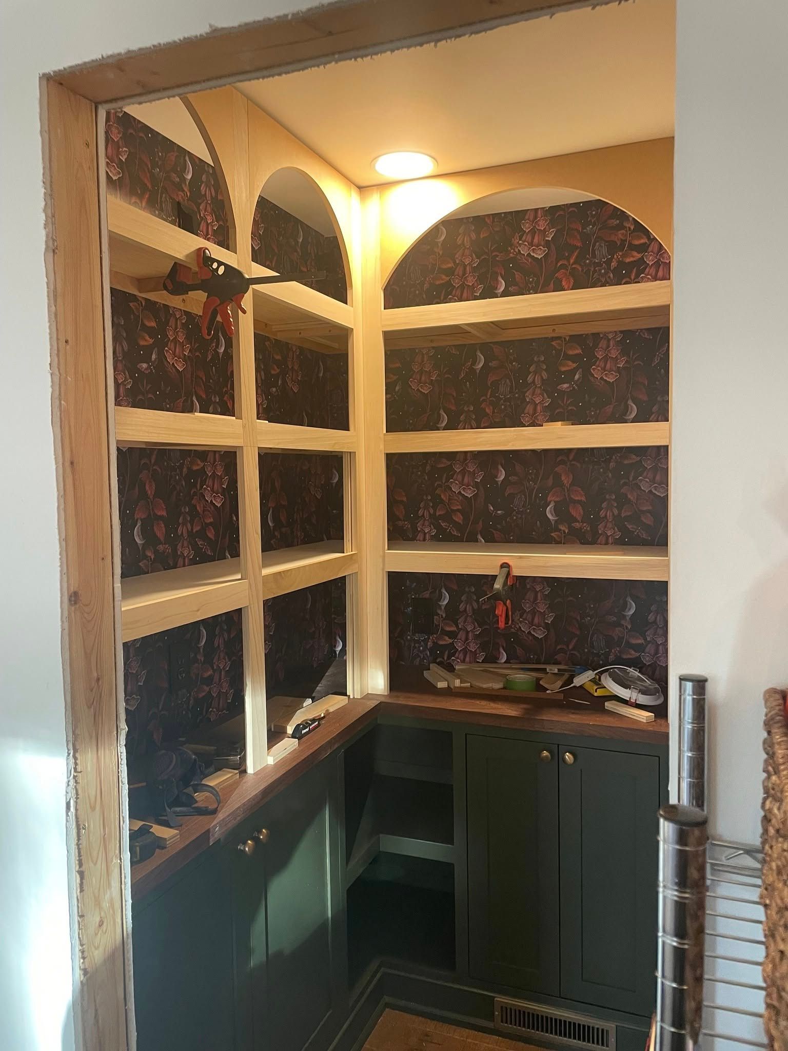 A walk-in pantry nook featuring dark green lower cabinets, wood shelves with arched details, and floral wallpaper.