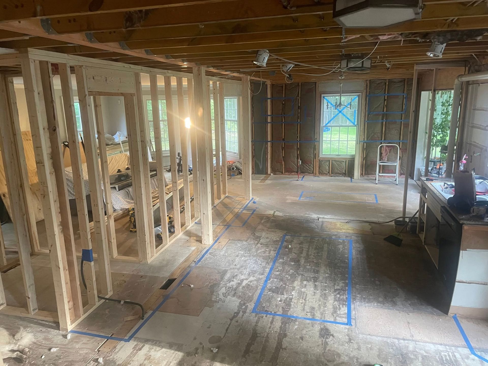 Interior of a house under renovation with wood-framed walls and exposed subflooring marked with blue painter's tape.