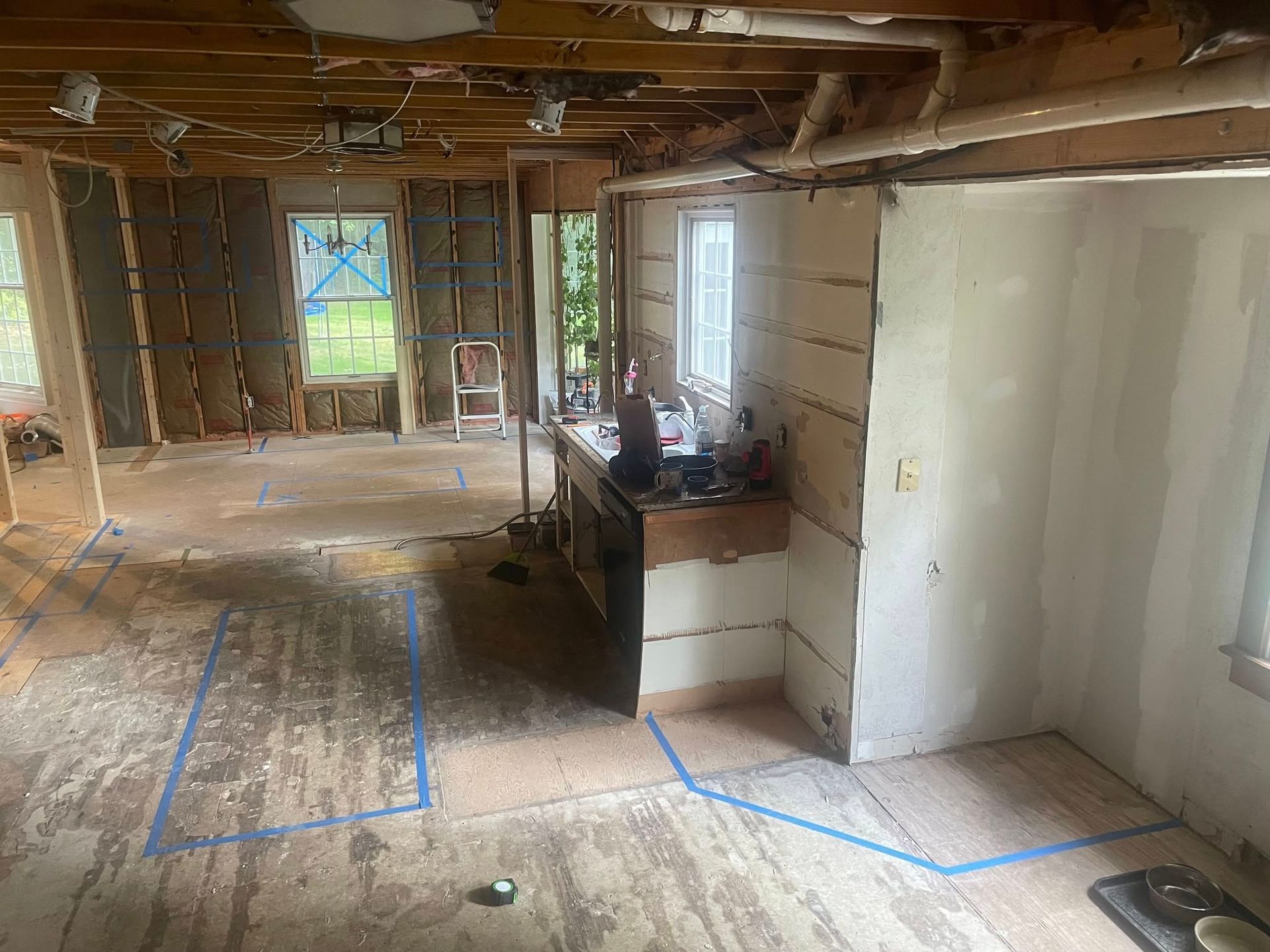 An interior room under renovation with exposed wall studs, blue painter's tape outlines on the floor, and a central sink.