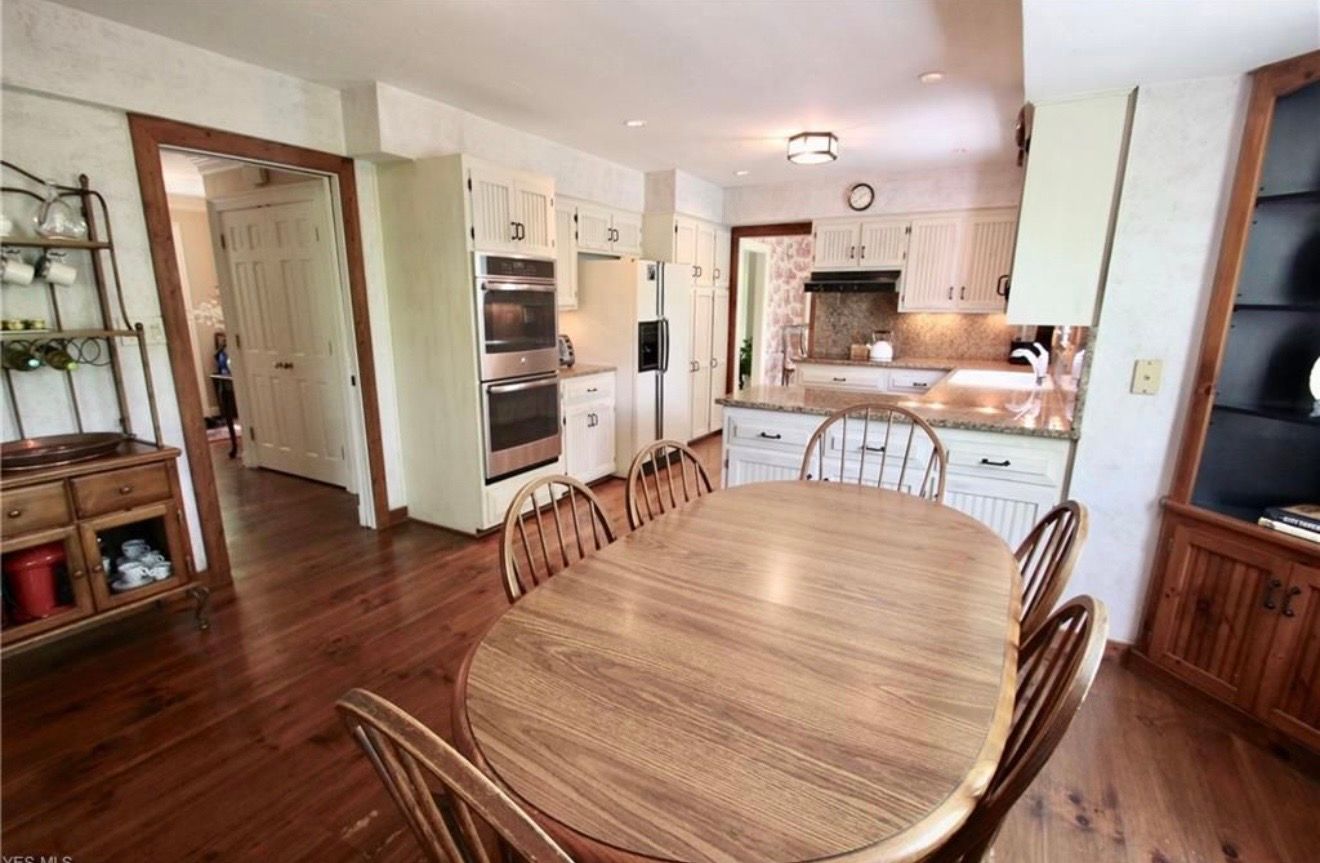 A dining area with a wooden oval table and chairs, adjacent to a kitchen with white cabinets and wood flooring.